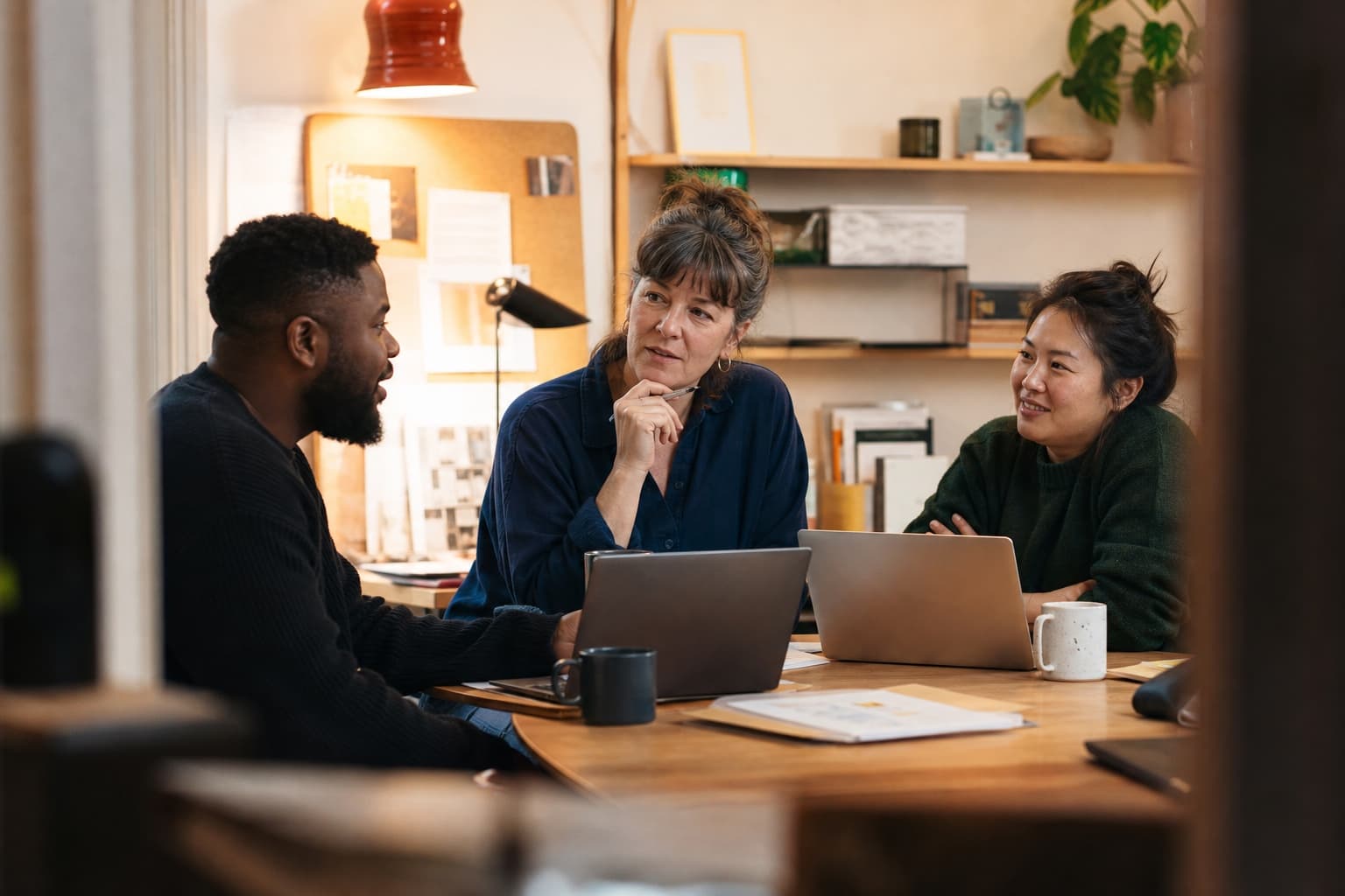 Three colleagues share ideas around laptops in a warm, small office.